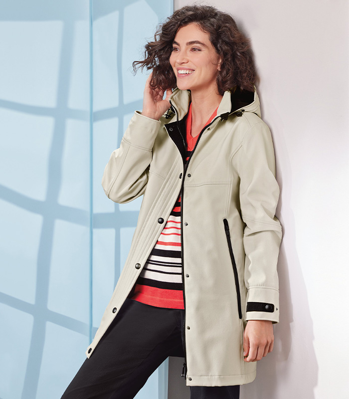 A happy woman leaning against a wall wearing a cream white all-weather coat with black buttons, lining and zip details, a coral, black and ivory cotton striped jumper and black casual trousers. 