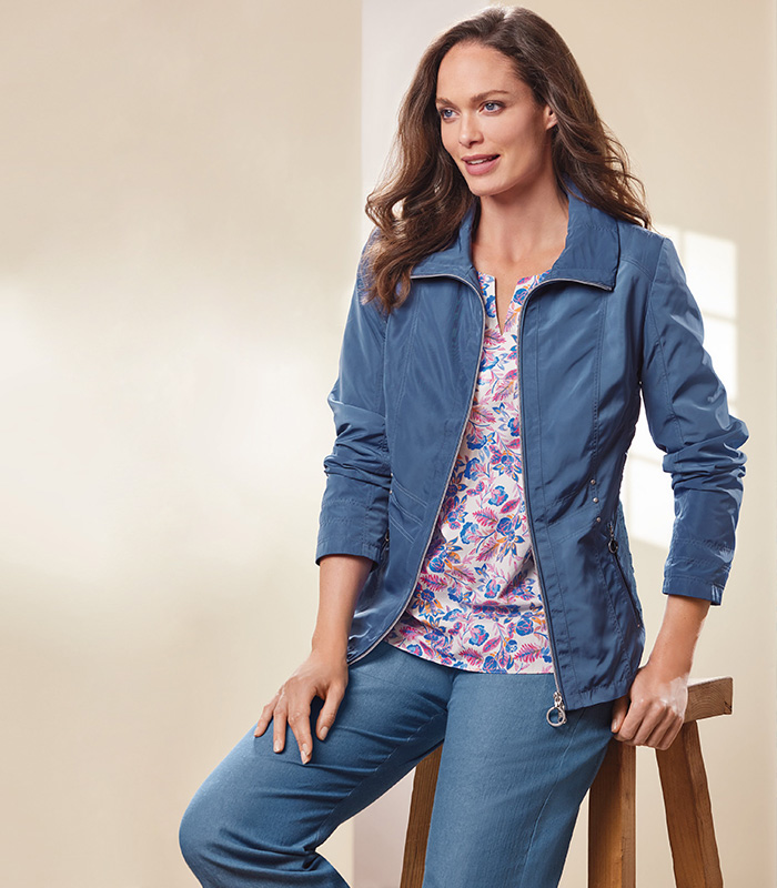 A woman perched on a stool smiling, wearing a blue casual coat along with a French floral printed tunic in tones of pink, blue and white.