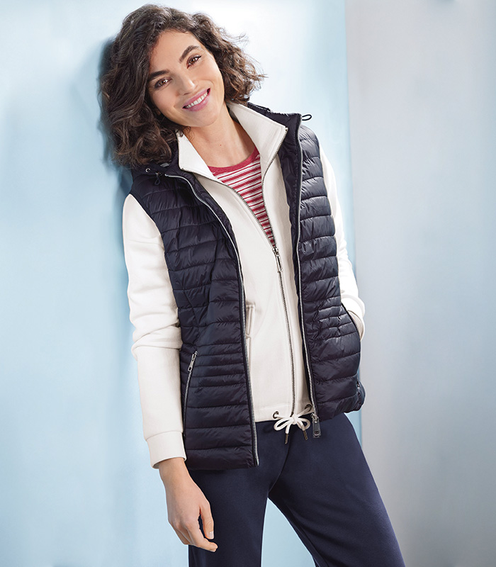 A woman leaning against a wall smiling, wearing a navy blue gilet, white jersey jacket, red and white striped jersey top and nay blue plain jersey trousers. 
