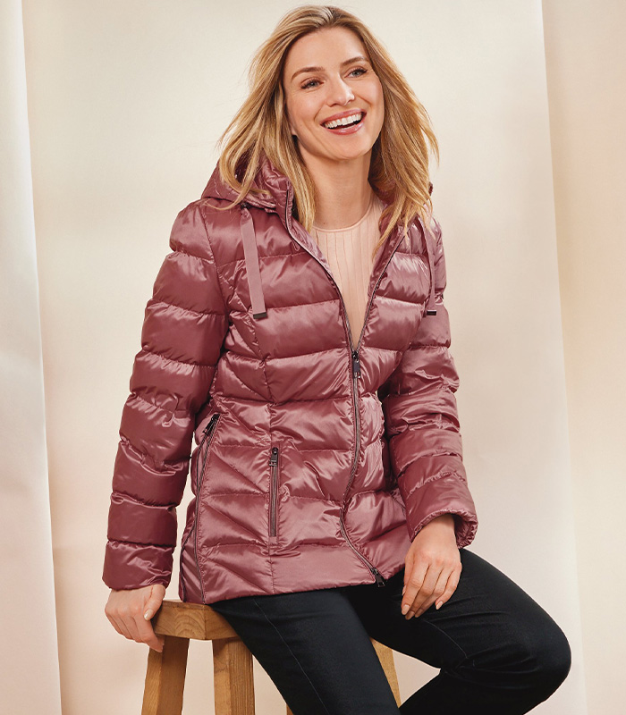 A woman sat on a stool smiling wearing a rose pink quilted coat, shell pink cotton-blend rib top and black casual trousers. 